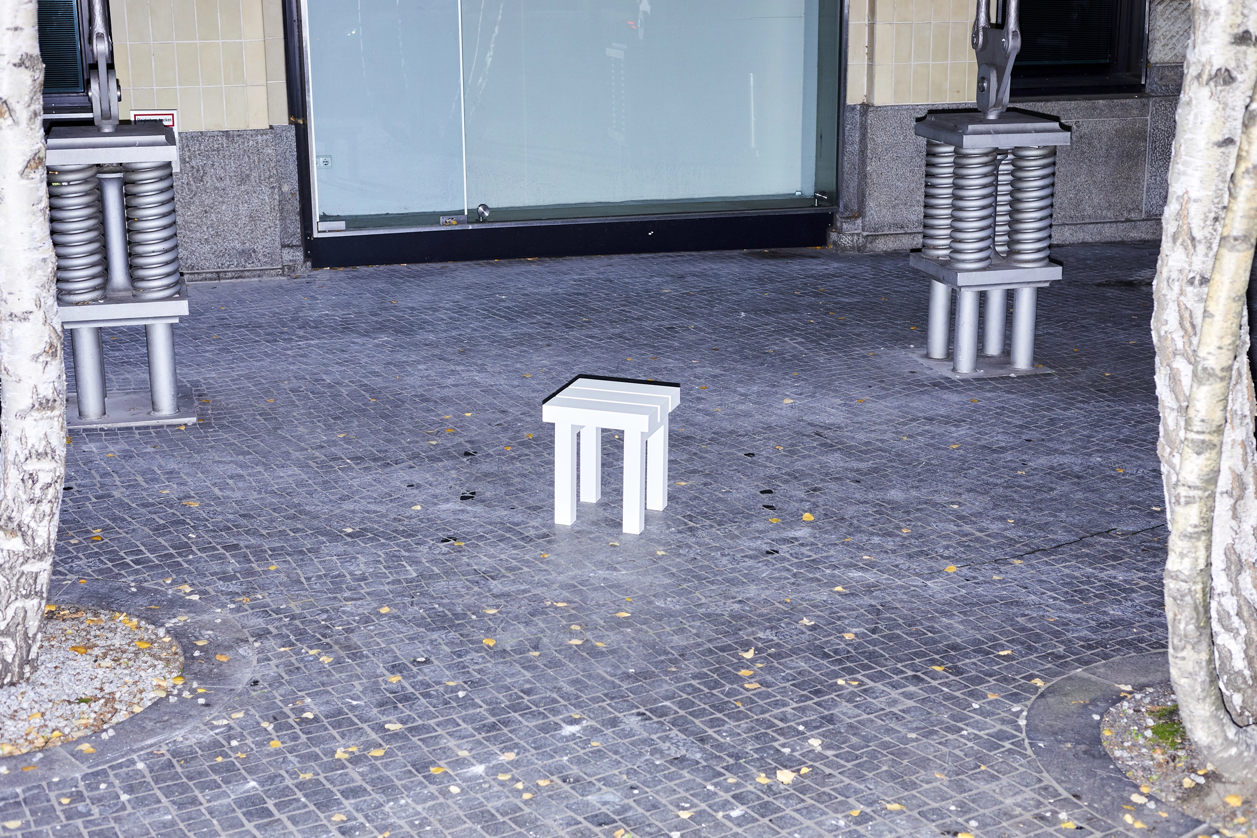 White aluminium stool from the furniture series Pretending It's Wood designed by ErtlundZull, photographed during the night on the sidewalk between two trees. ErtlundZull